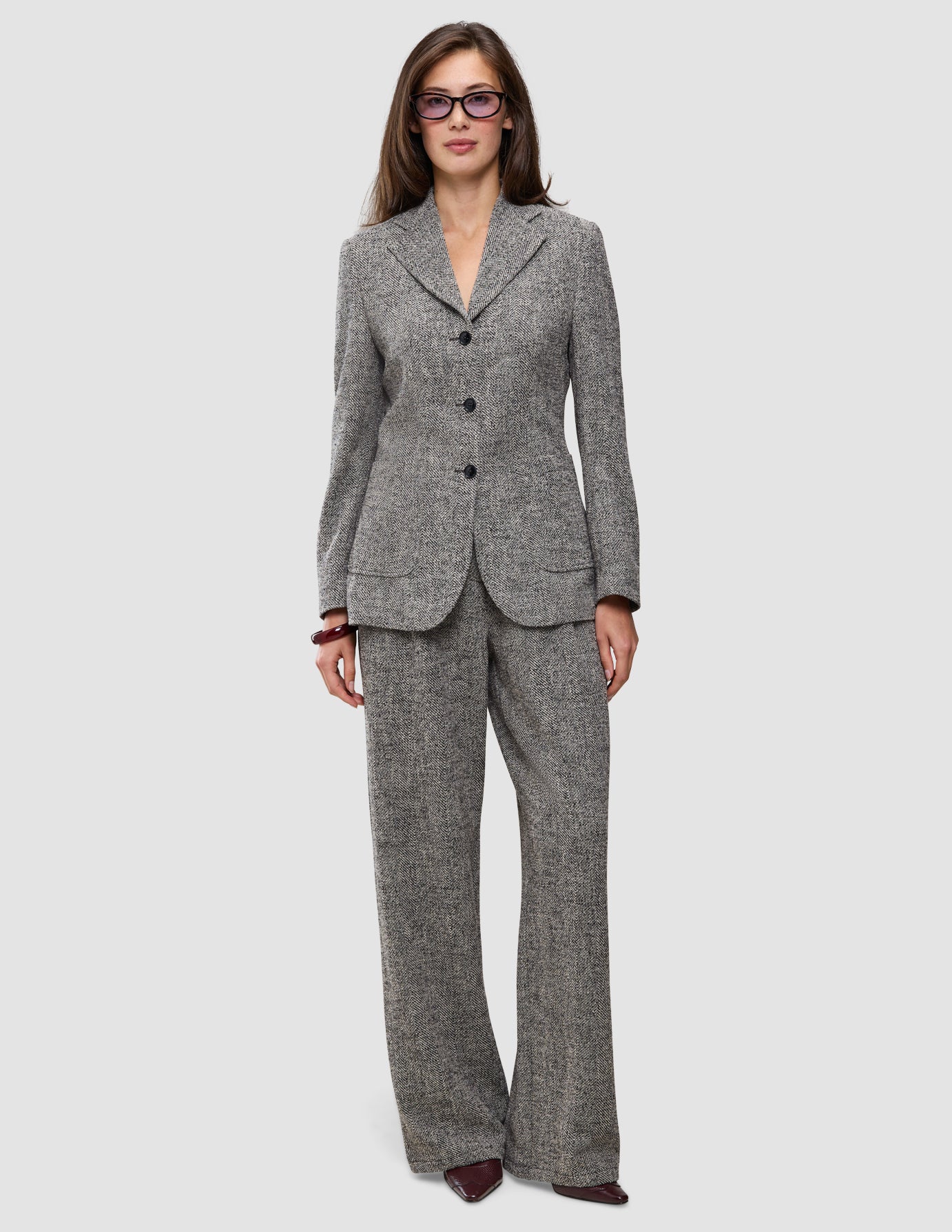 Eine Frau trägt einen grauen, strukturierten Women Ashryn Pants Wide Domino Herringbone-Anzug aus Bouclé-Stoff mit Fischgrätenmuster, bestehend aus einem Blazer mit drei Knöpfen und einer hoch taillierten Hose mit weitem Bein.